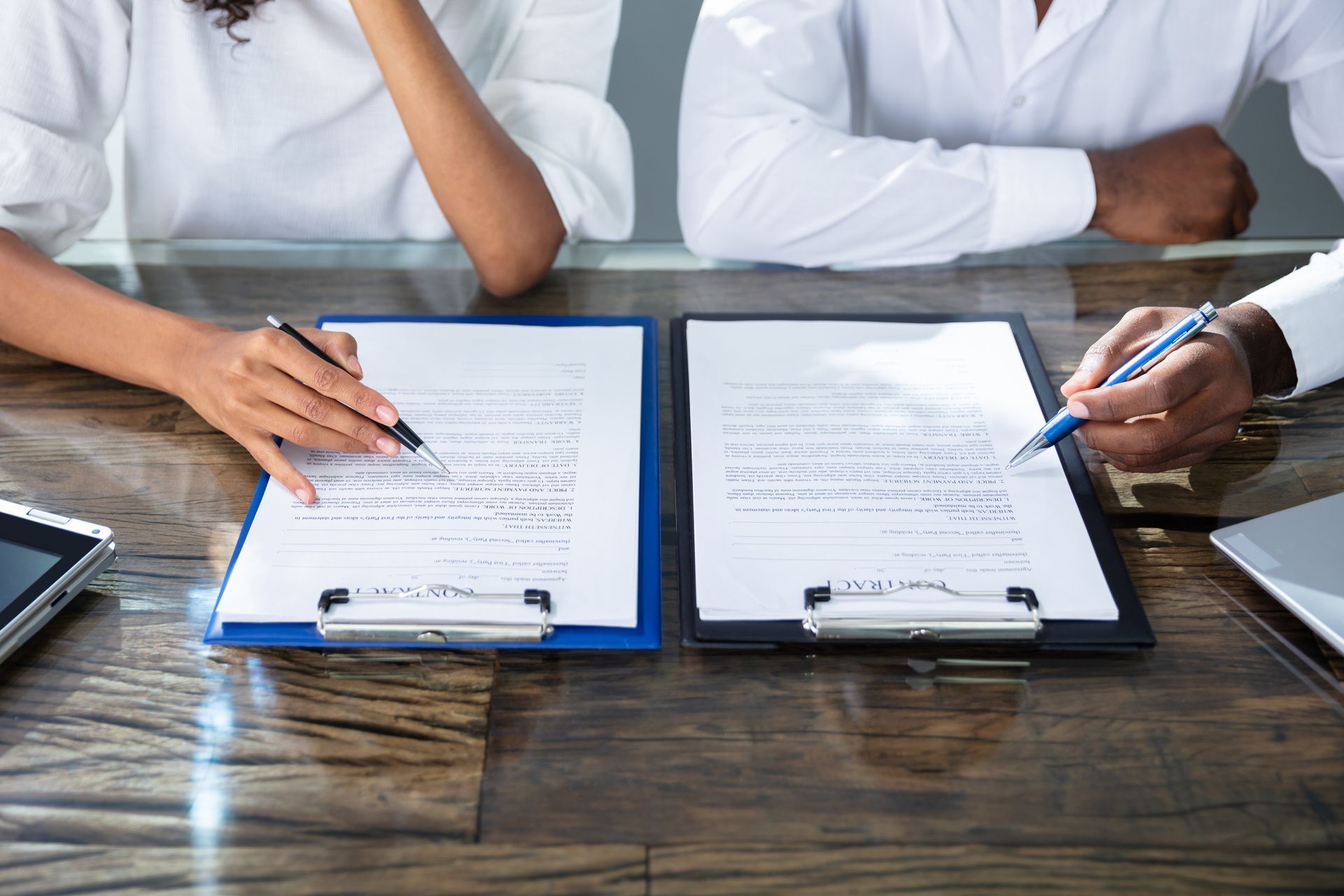 Two people in white shirts use pens to review and sign documents on clipboards placed on a polished wooden table.