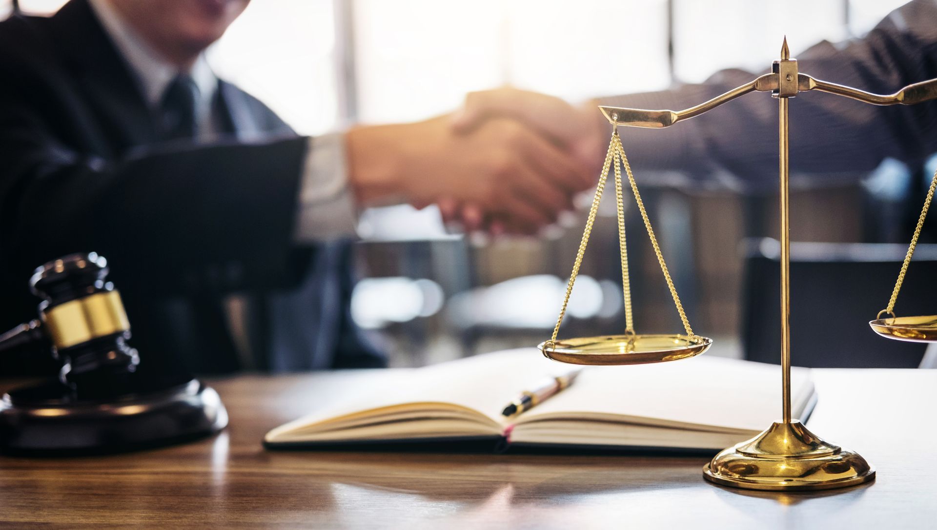 A judge's gavel and gold scales of justice on a desk in front of two professionals shaking hands in a legal setting.