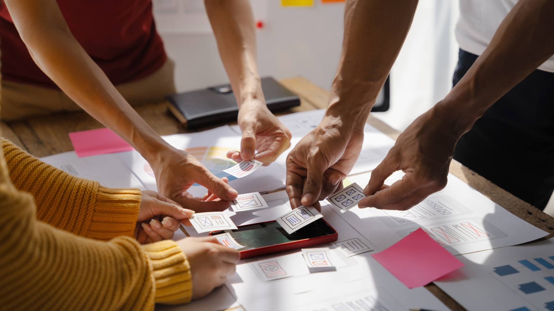 People’s hands point to a smartphone on a table with papers and sticky notes during a brainstorming session