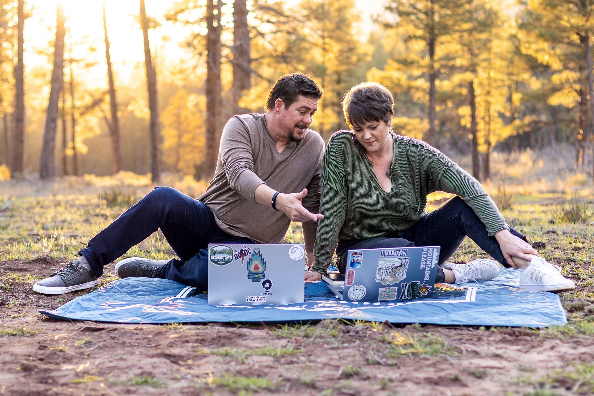 Two people sitting on a blanket outdoors, using laptops in a sunny park.