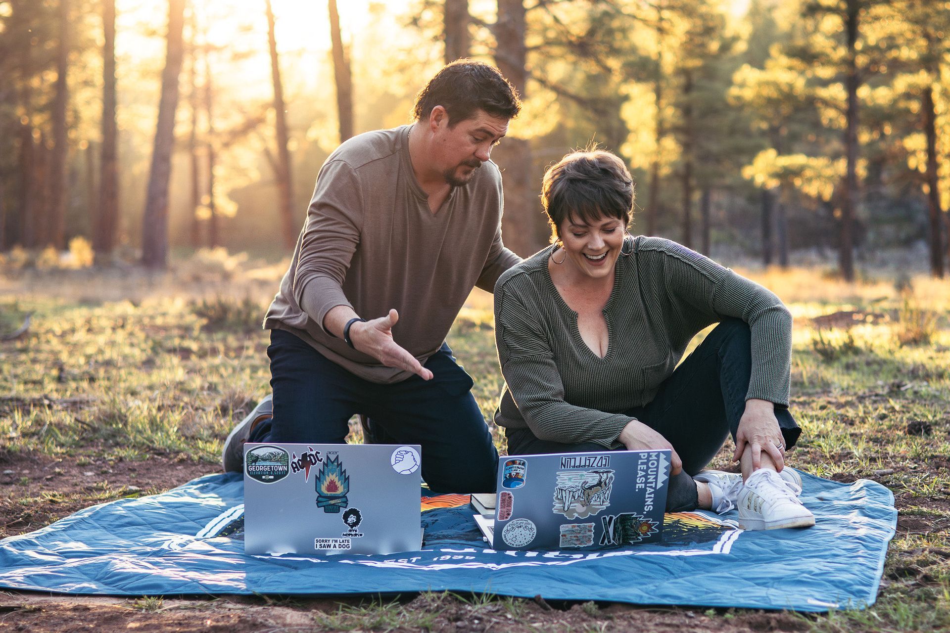 Two people sitting on a blanket outdoors, smiling at laptops in a sunlit wooded park.