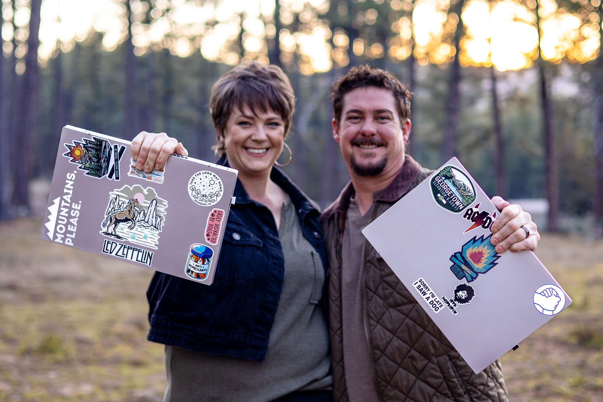 Two people smiling outdoors at sunset, each holding up a decorated laptop or binder.