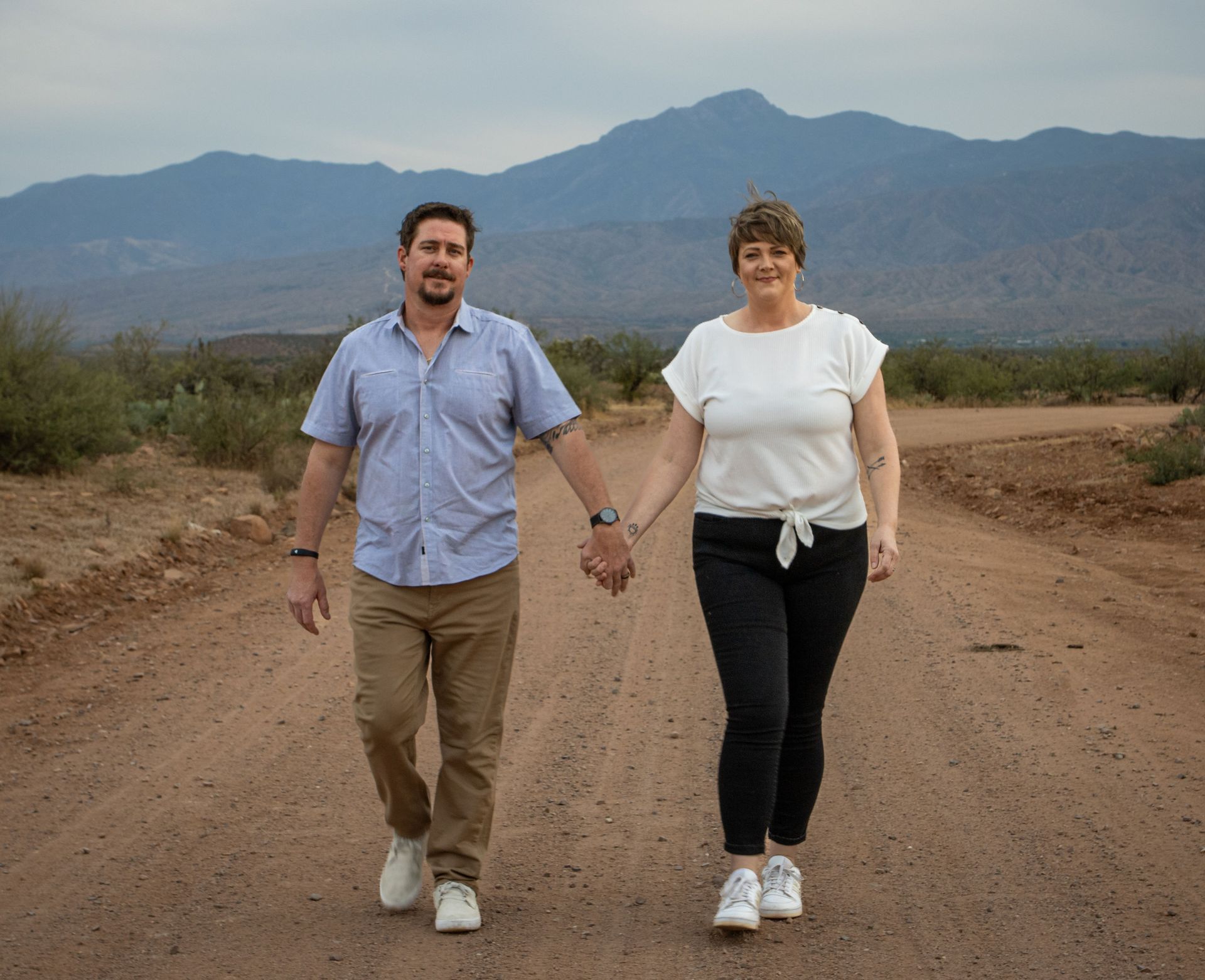 Two people holding hands and walking on a dirt road with mountains in the background
