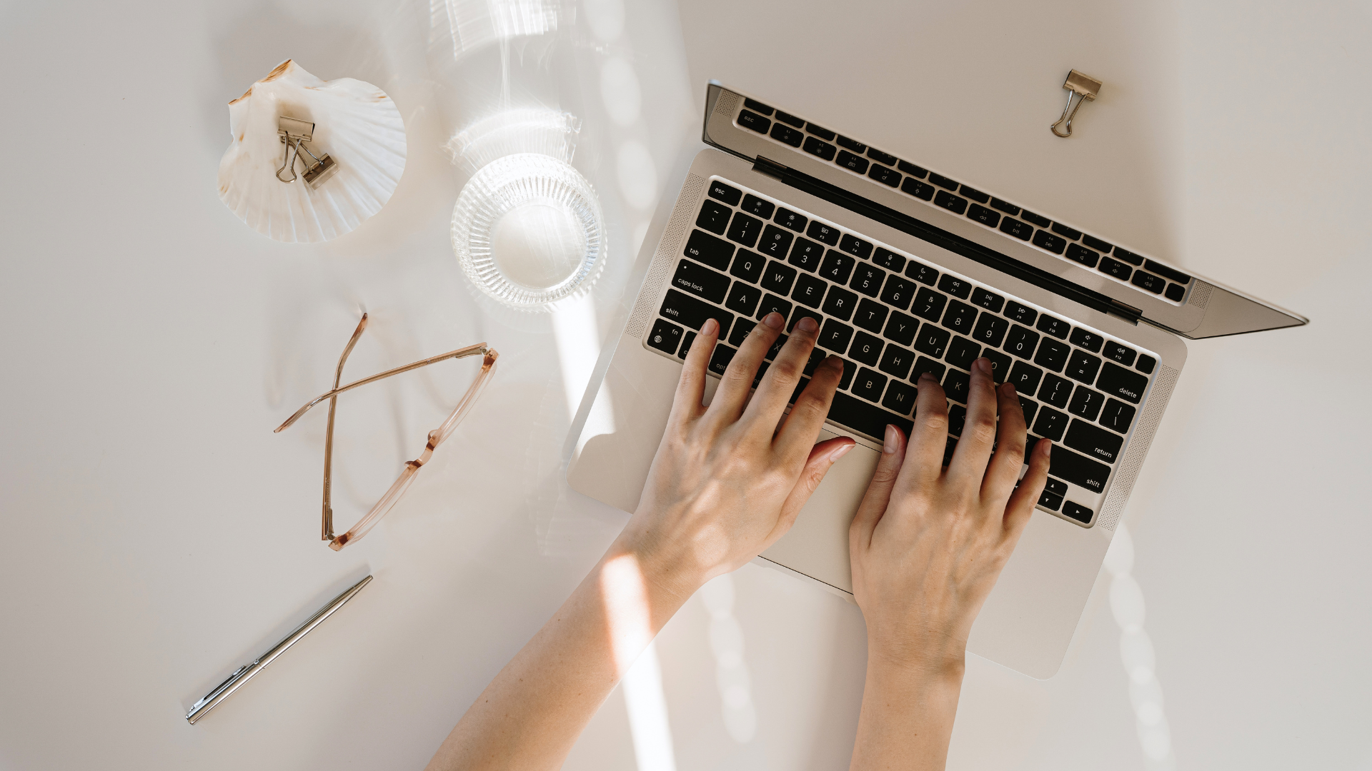 Hands typing on a laptop at a bright desk with coffee, glasses, and a pen.