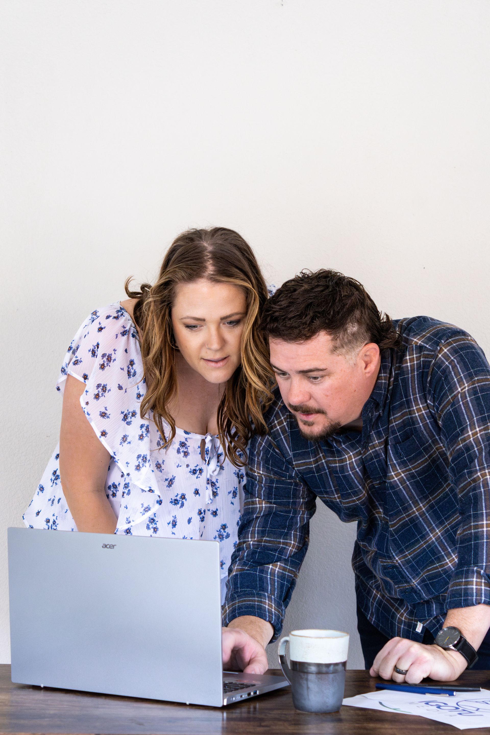 Two coworkers leaning over a laptop, reviewing notes at a table with coffee cups.