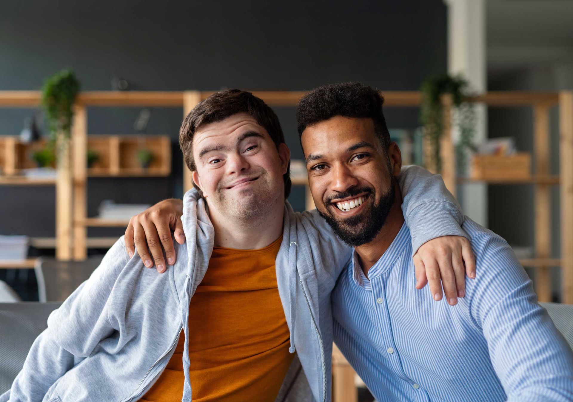 Young man with Down syndrome and his tutor with arms around him looking at the camera indoors. Young man with Down syndrome and his tutor with arms around him looking at the camera indoors.