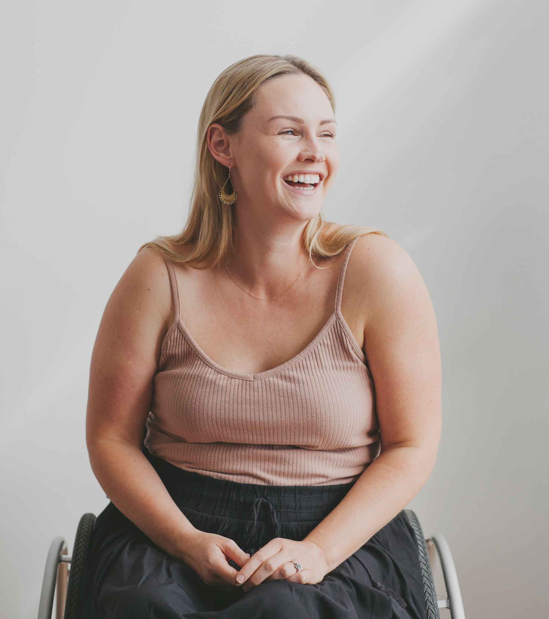 Woman in a wheelchair smiles, looking to the side. She wears a tan tank top and black skirt against a white wall.