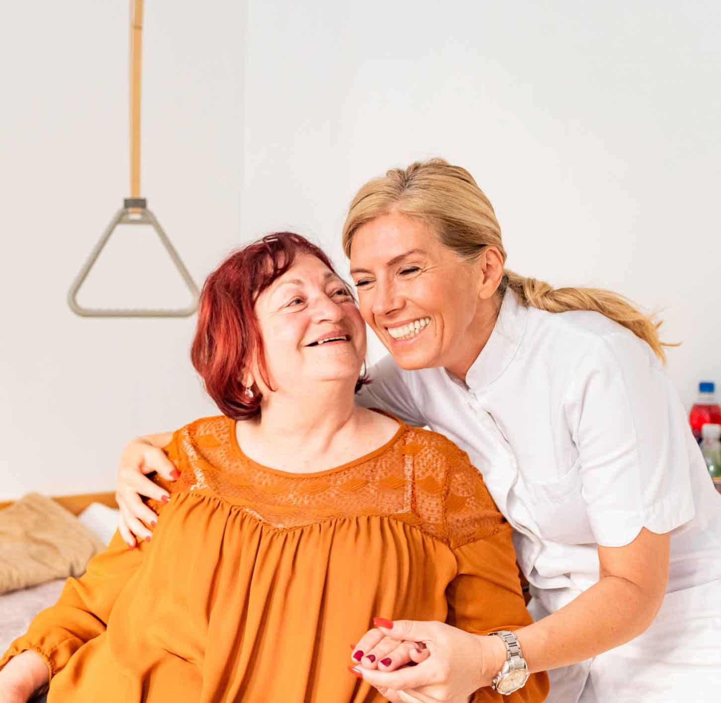 A smiling caregiver embraces an older woman with red hair, offering support in a home-care setting. A smiling caregiver embraces an older woman with red hair, offering support in a home-care setting.