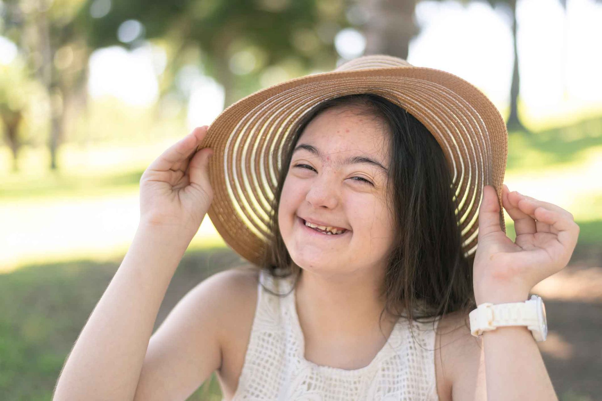 Young woman with Down syndrome smiling, wearing a sun hat in a park setting.