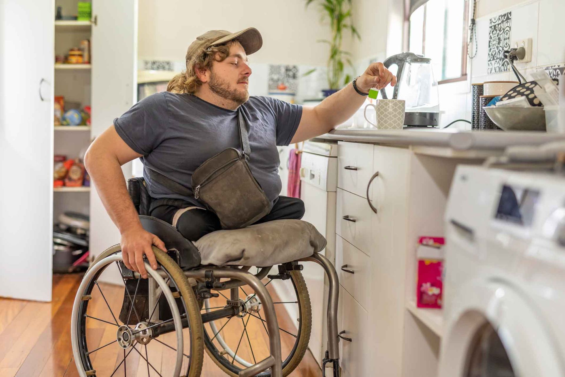 A man in a wheelchair reaches toward a window in a kitchen. He wears a cap and blue shirt. White cabinets and appliances are visible.