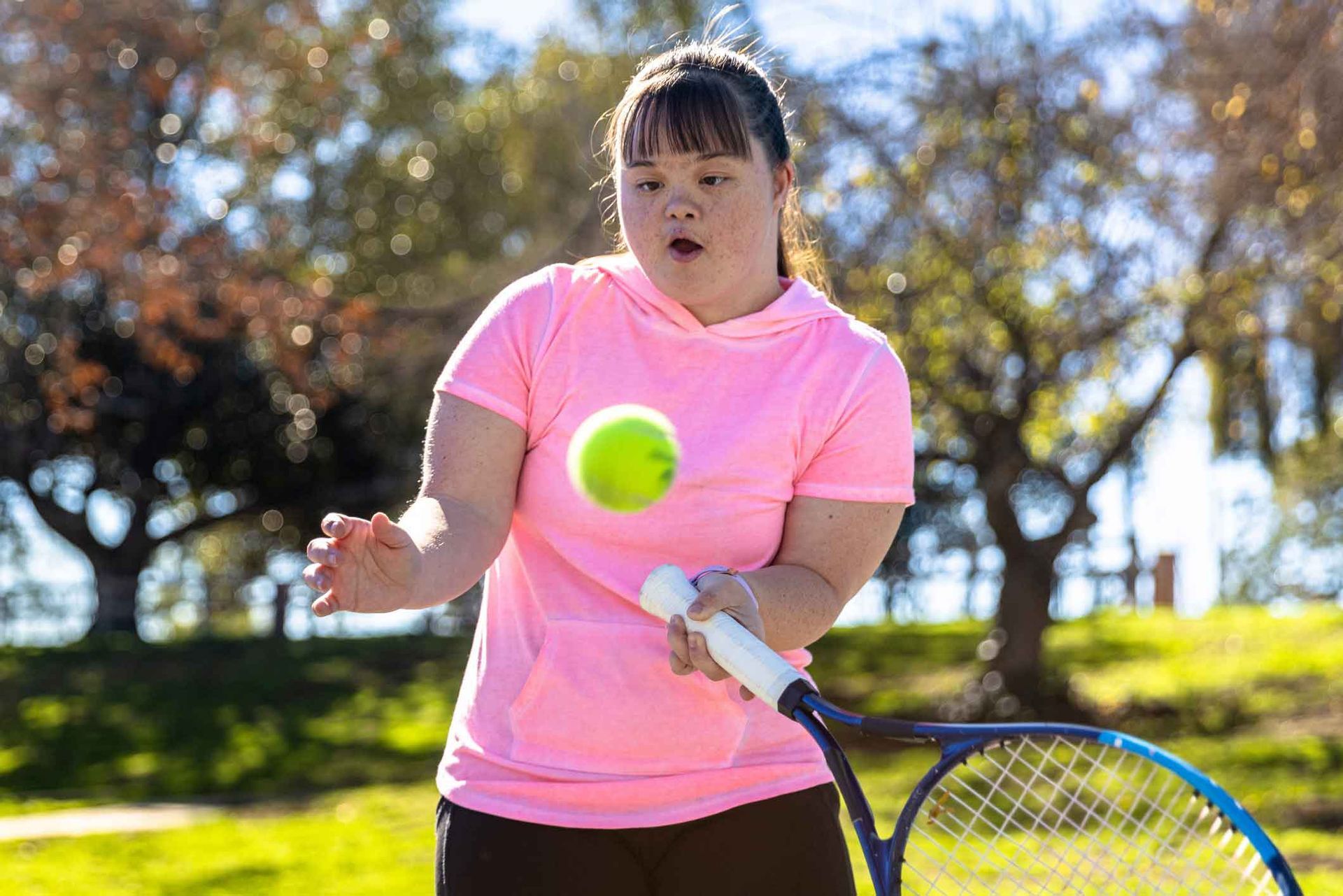 A young woman with Down syndrome in a pink shirt plays tennis outdoors. She focuses on the ball, ready to hit it with her racquet. A young woman with Down syndrome in a pink shirt plays tennis outdoors. She focuses on the ball, ready to hit it with her racquet.