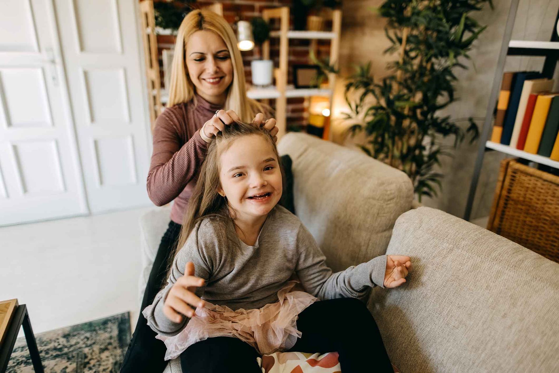 Woman braiding a young girl's hair on a sofa in a living room. Both are smiling; the girl holds a toy. Woman braiding a young girl's hair on a sofa in a living room. Both are smiling; the girl holds a toy.