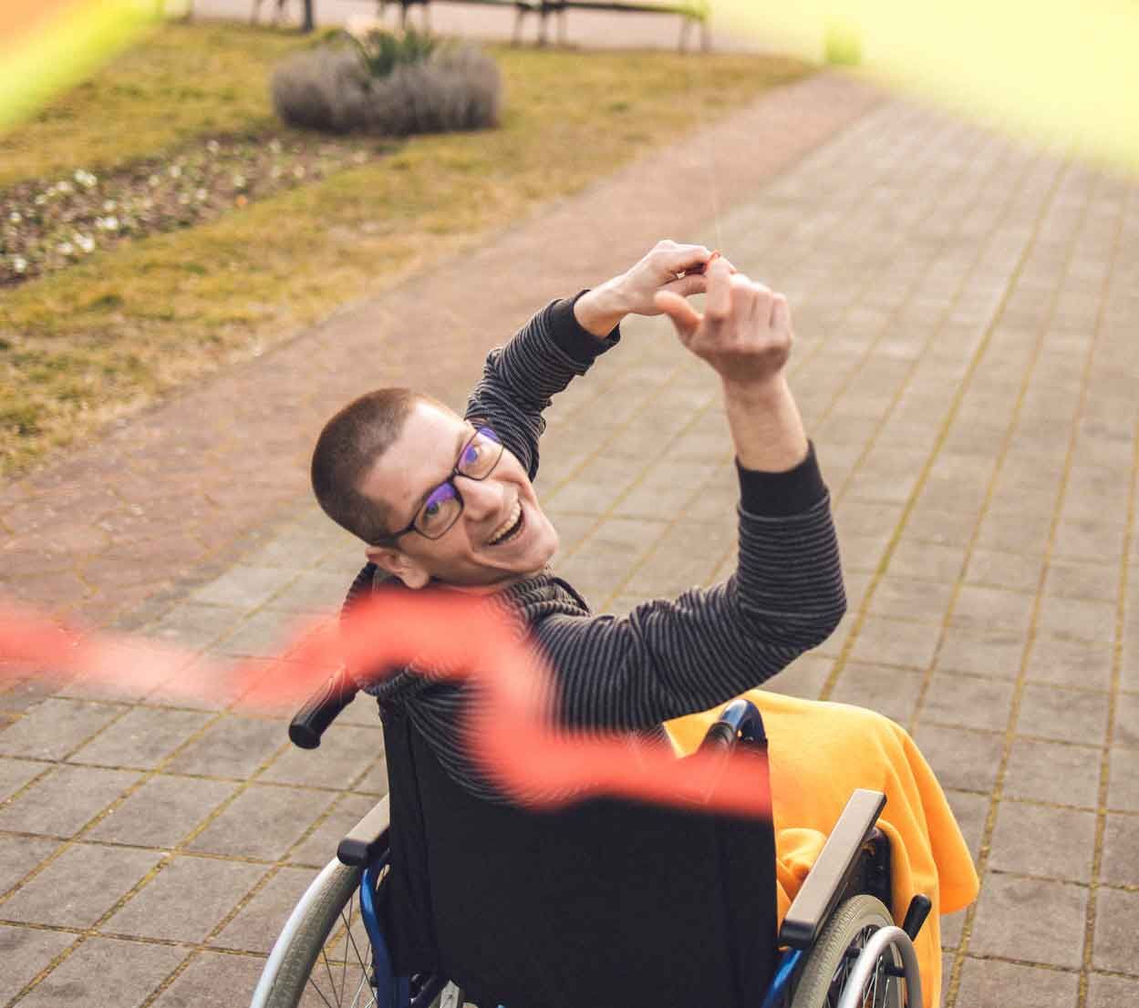 Smiling person in a wheelchair outdoors, raising their arms. They wear glasses and a dark jacket, orange blanket draped on chair. Smiling person in a wheelchair outdoors, raising their arms. They wear glasses and a dark jacket, orange blanket draped on chair.