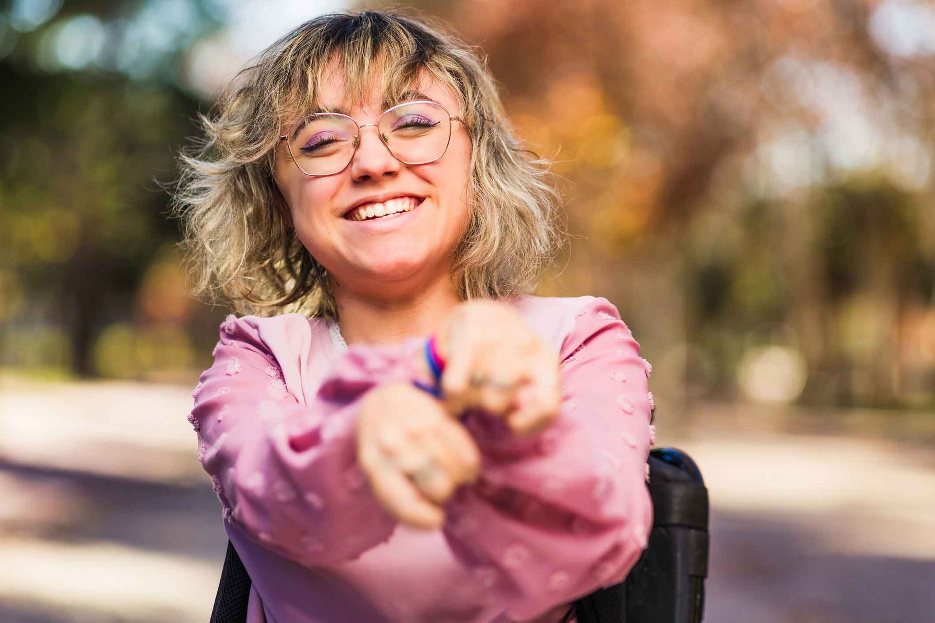 Woman in a wheelchair smiles, holding up hands. She has short blonde hair, glasses, and a pink shirt, in a park.