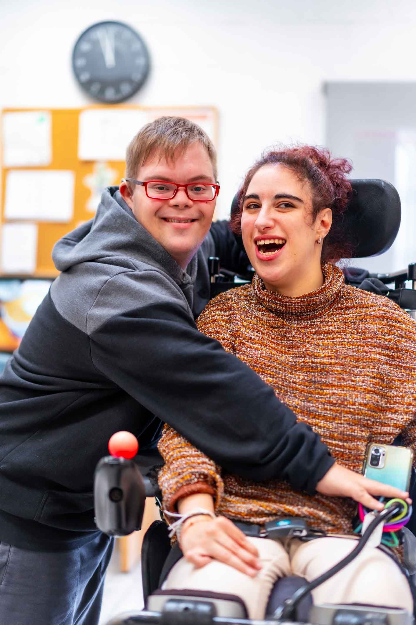 Man with Down syndrome embraces a woman in a wheelchair; both are smiling and laughing in a brightly lit room. Man with Down syndrome embraces a woman in a wheelchair; both are smiling and laughing in a brightly lit room.