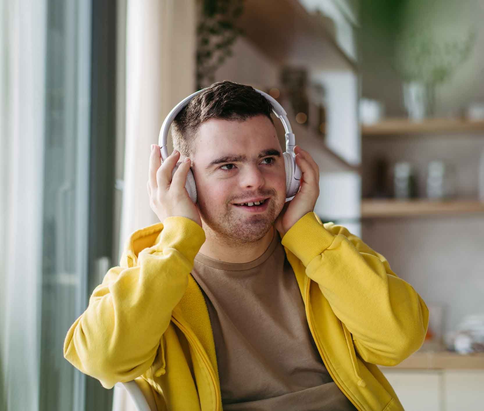 A man with Down syndrome wearing a yellow jacket and headphones, smiling while looking to the right. A man with Down syndrome wearing a yellow jacket and headphones, smiling while looking to the right.