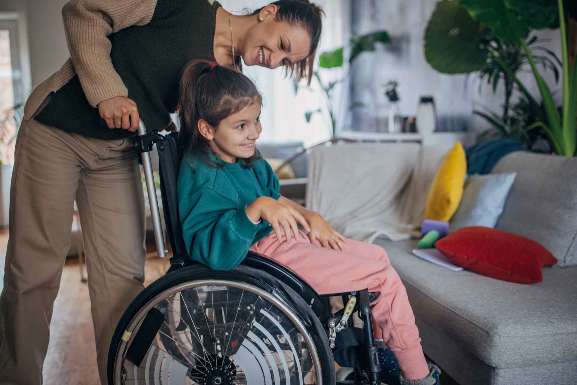 A woman smiles at a young girl in a wheelchair. The girl is sitting indoors, looking to the side, as the woman pushes her. A woman smiles at a young girl in a wheelchair. The girl is sitting indoors, looking to the side, as the woman pushes her.
