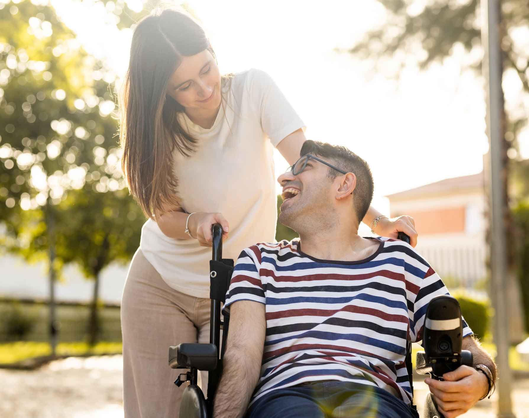 Woman smiles at a laughing man in a wheelchair, pushing him outdoors in sunlight.
