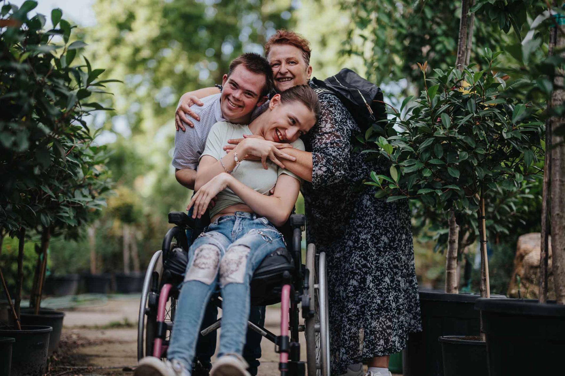 Three smiling people hugging: a woman in a wheelchair, a man, and another woman, in a plant nursery. Three smiling people hugging: a woman in a wheelchair, a man, and another woman, in a plant nursery.