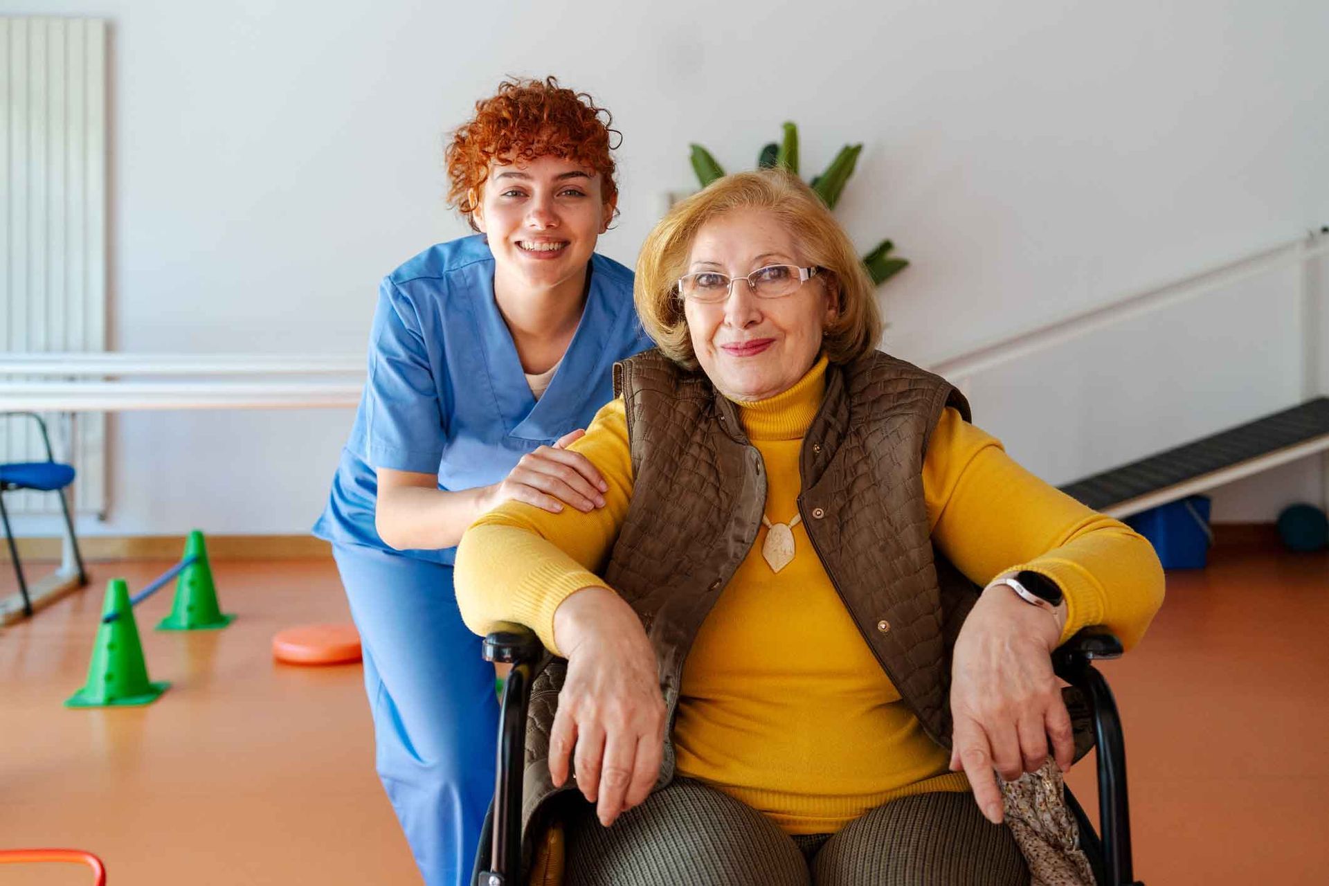 A smiling caregiver in blue scrubs assists a senior woman in a wheelchair wearing a yellow sweater and vest. They are in a therapy room. A smiling caregiver in blue scrubs assists a senior woman in a wheelchair wearing a yellow sweater and vest. They are in a therapy room.