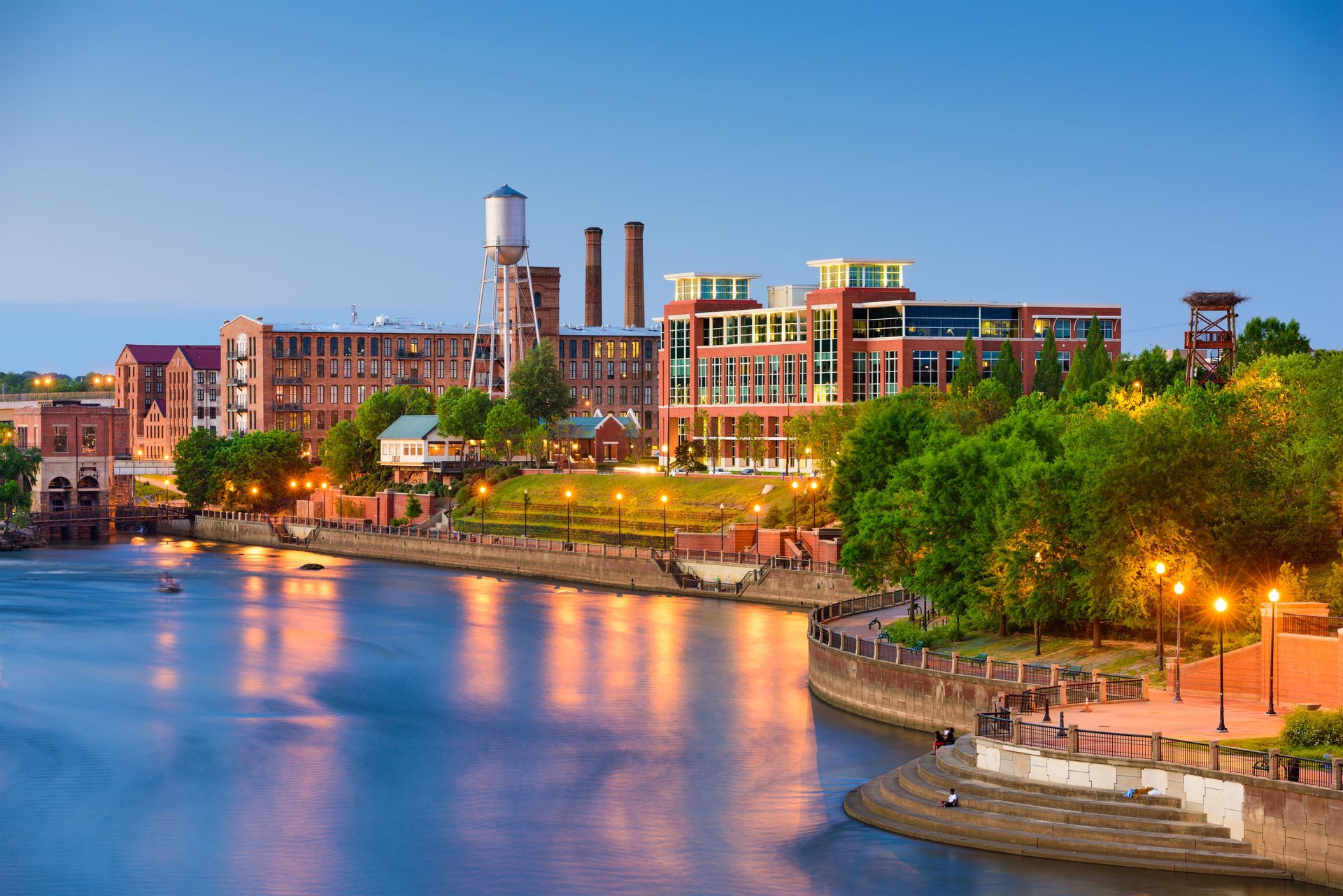 City waterfront at dusk with lit buildings, smokestacks, trees, and calm river reflections