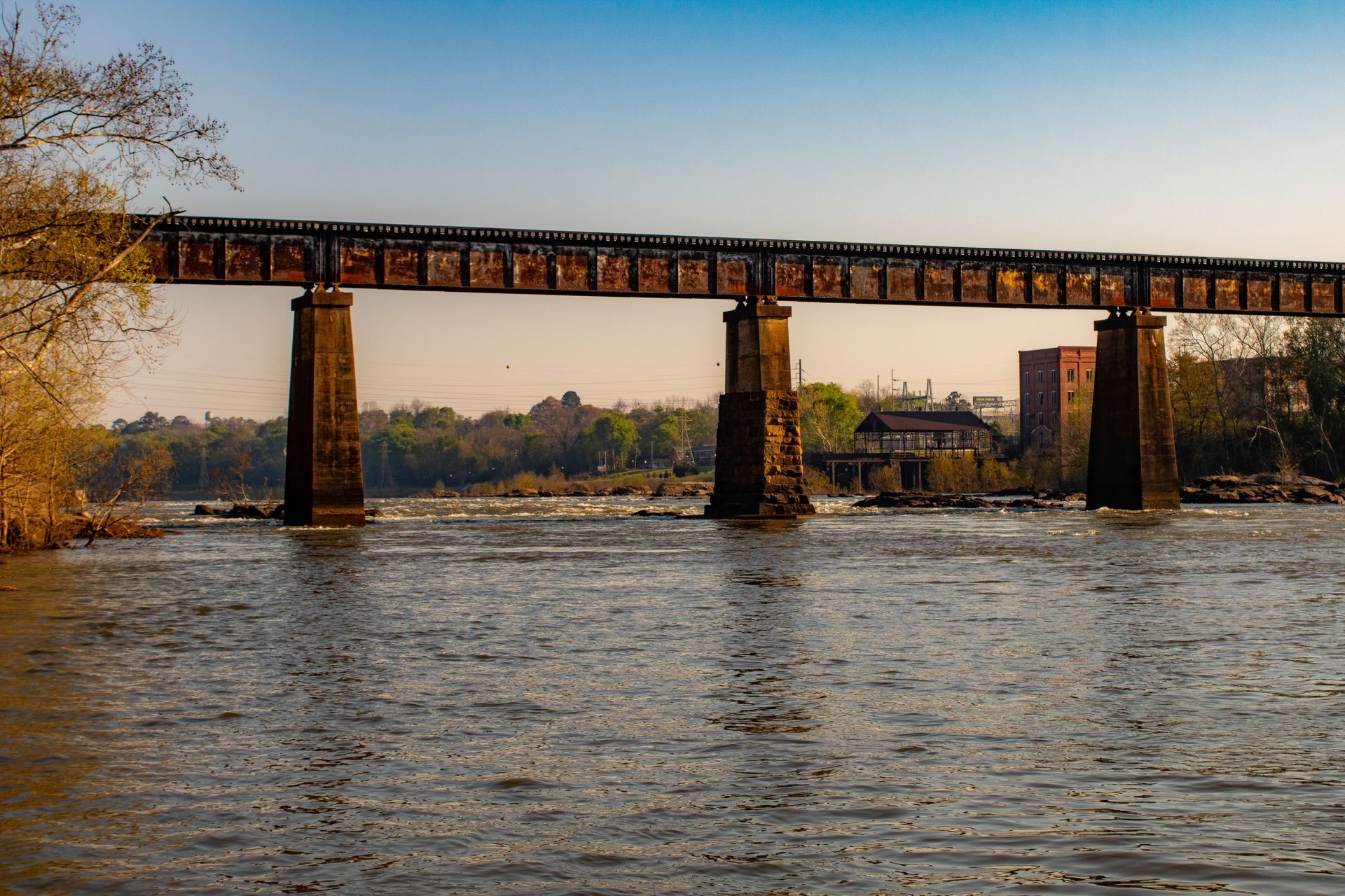 A bridge spans a wide river at sunset, with trees and buildings in the background.