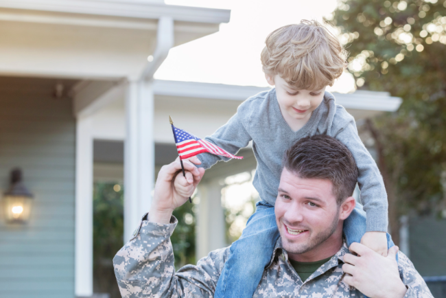 Soldier carrying a child on his shoulders while the child waves a small American flag outdoors