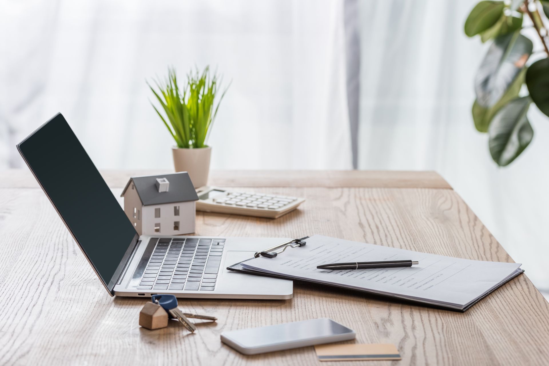 Laptop and notebook on a wooden desk with a small house model, plant, keys, and smartphone nearby