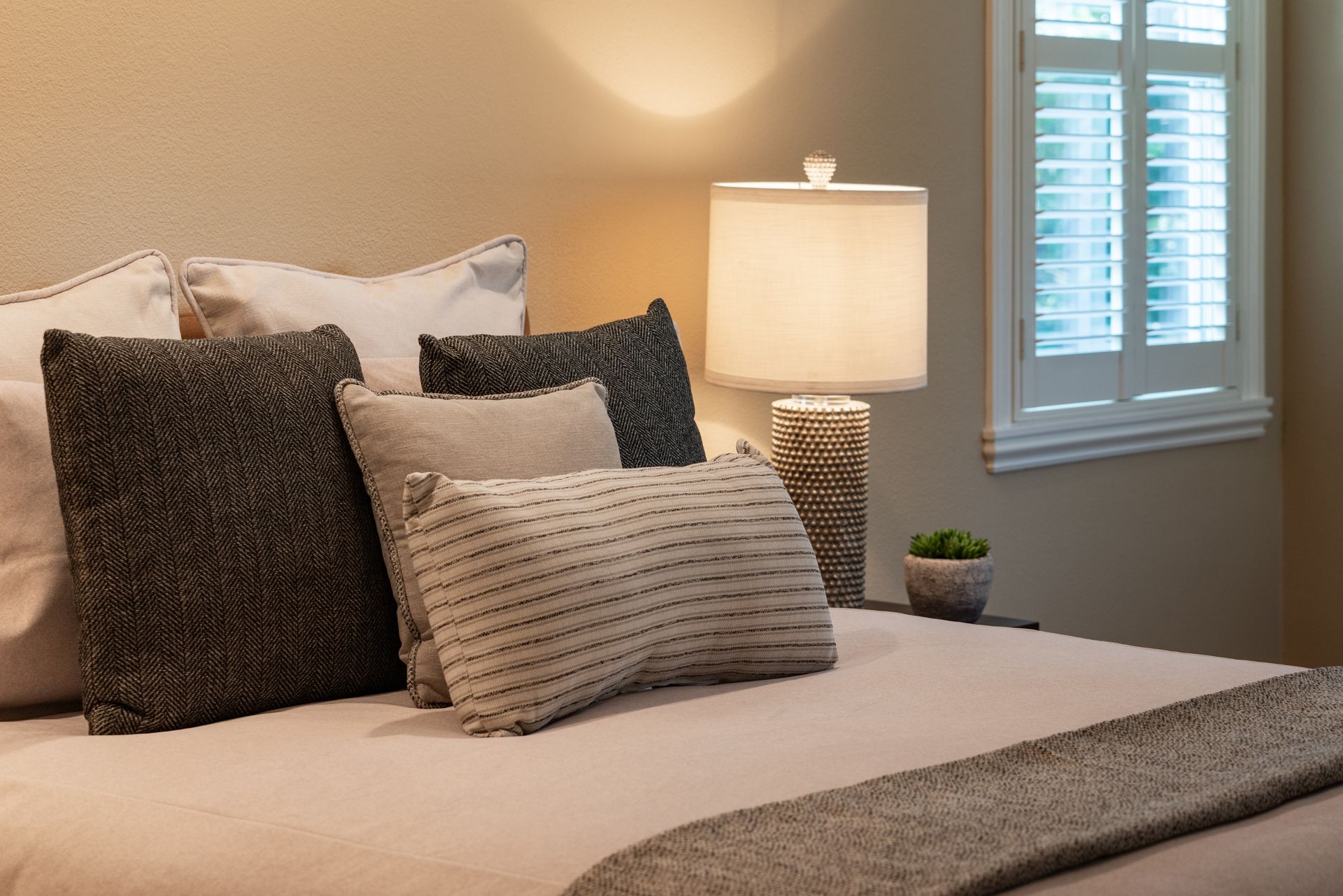 Cozy bedroom with beige bedding, patterned pillows, a lit lamp, and white window shutters