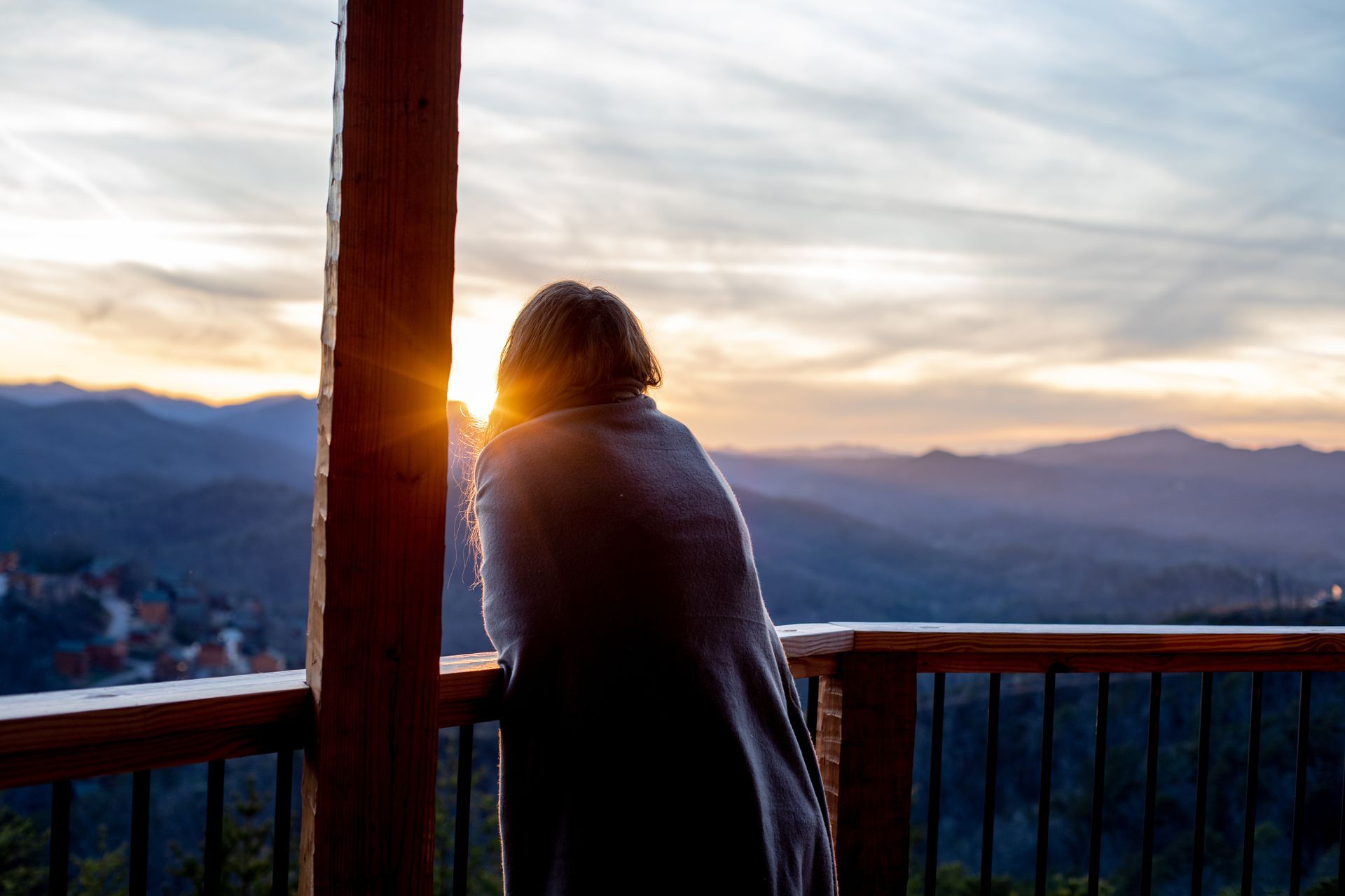 A woman is standing on a balcony looking at the sunset over the mountains.
