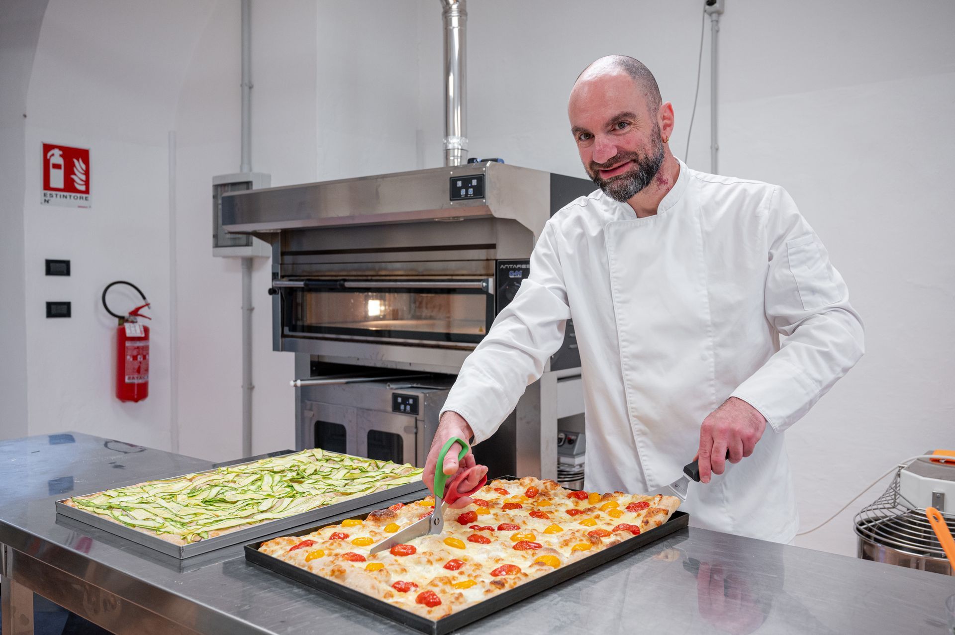 In una luminosa cucina professionale, lo chef sistema le pizze sui vassoi, con un forno sullo sfondo.
