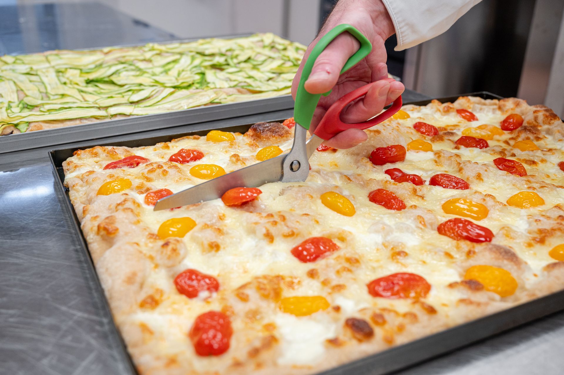 Tagliare a mano una pizza con peperoni e pomodoro su una teglia da forno con forbici dal manico verde.