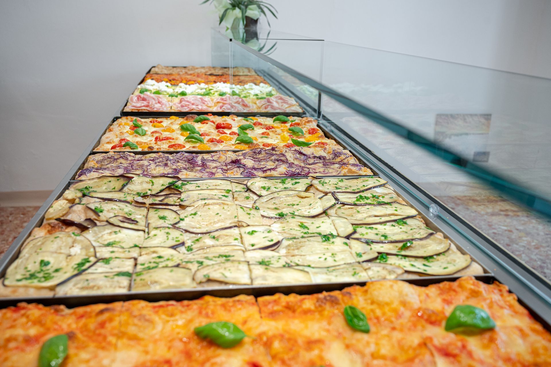Un buffet con vassoi di focacce colorate e piatti con verdure, protetto da una barriera di vetro parasputi.