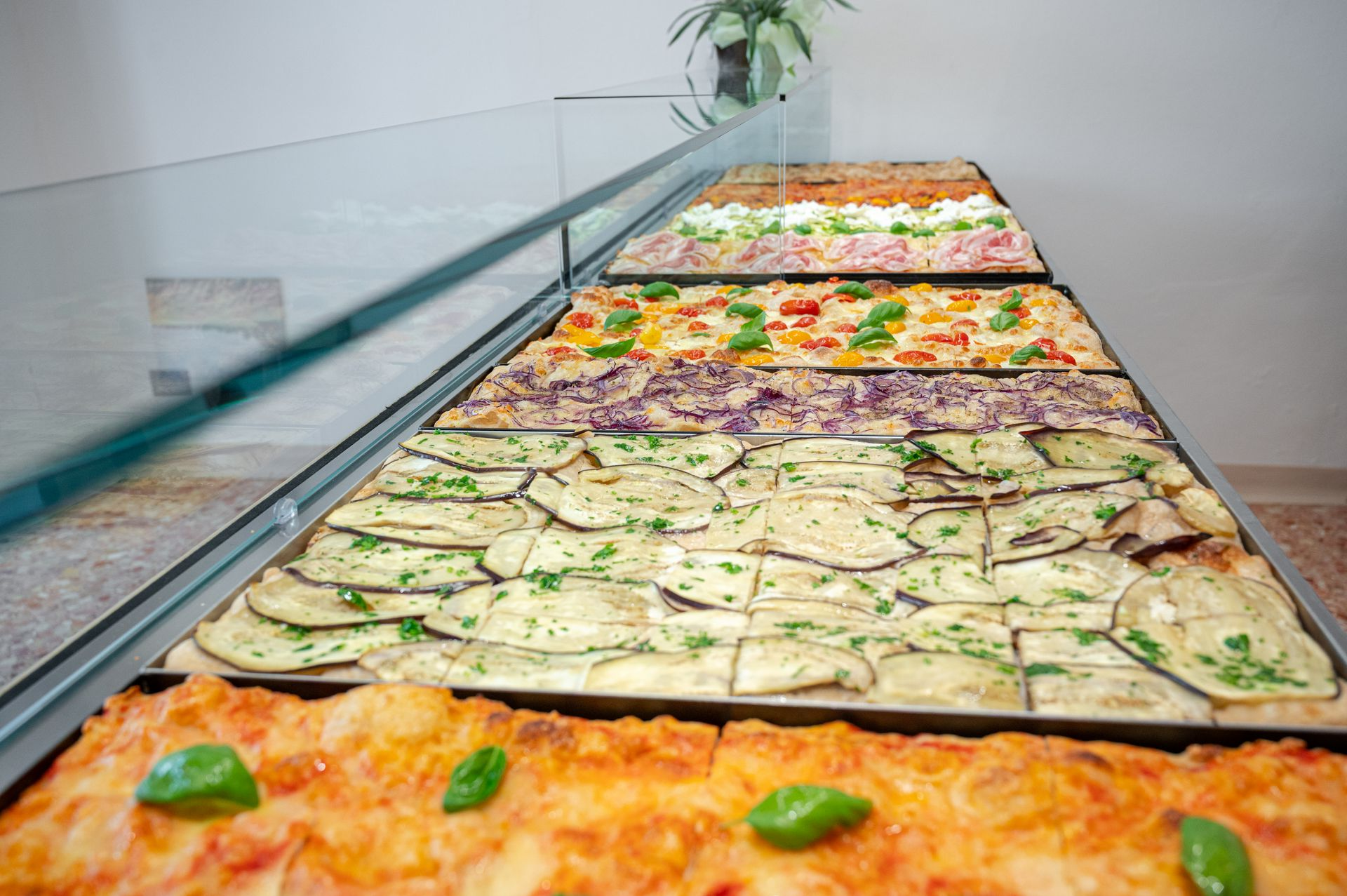 Lungo buffet di pizze rettangolari assortite su un bancone di vetro in una luminosa caffetteria.
