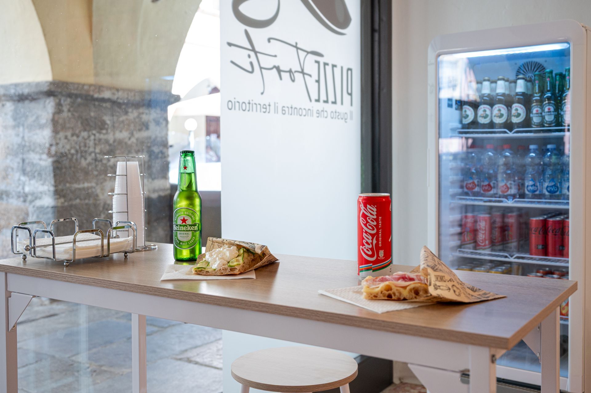 Un tavolo con snack, bottiglia di birra, lattina di bibita e pizza in un bar luminoso vicino a un frigorifero.