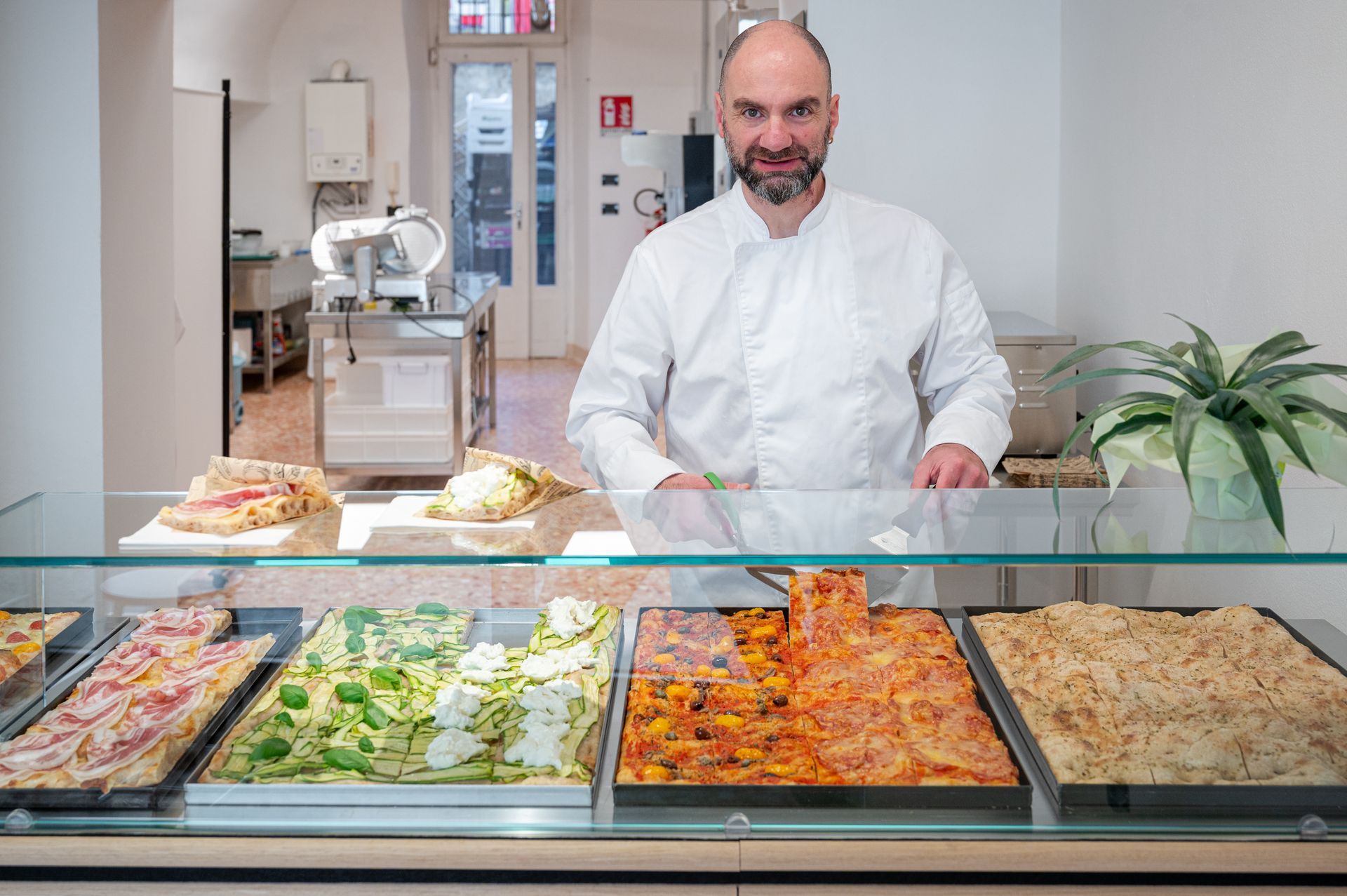 Uno chef dietro il bancone di una gastronomia, con vassoi di cibi pronti e pasticcini in un negozio luminoso.