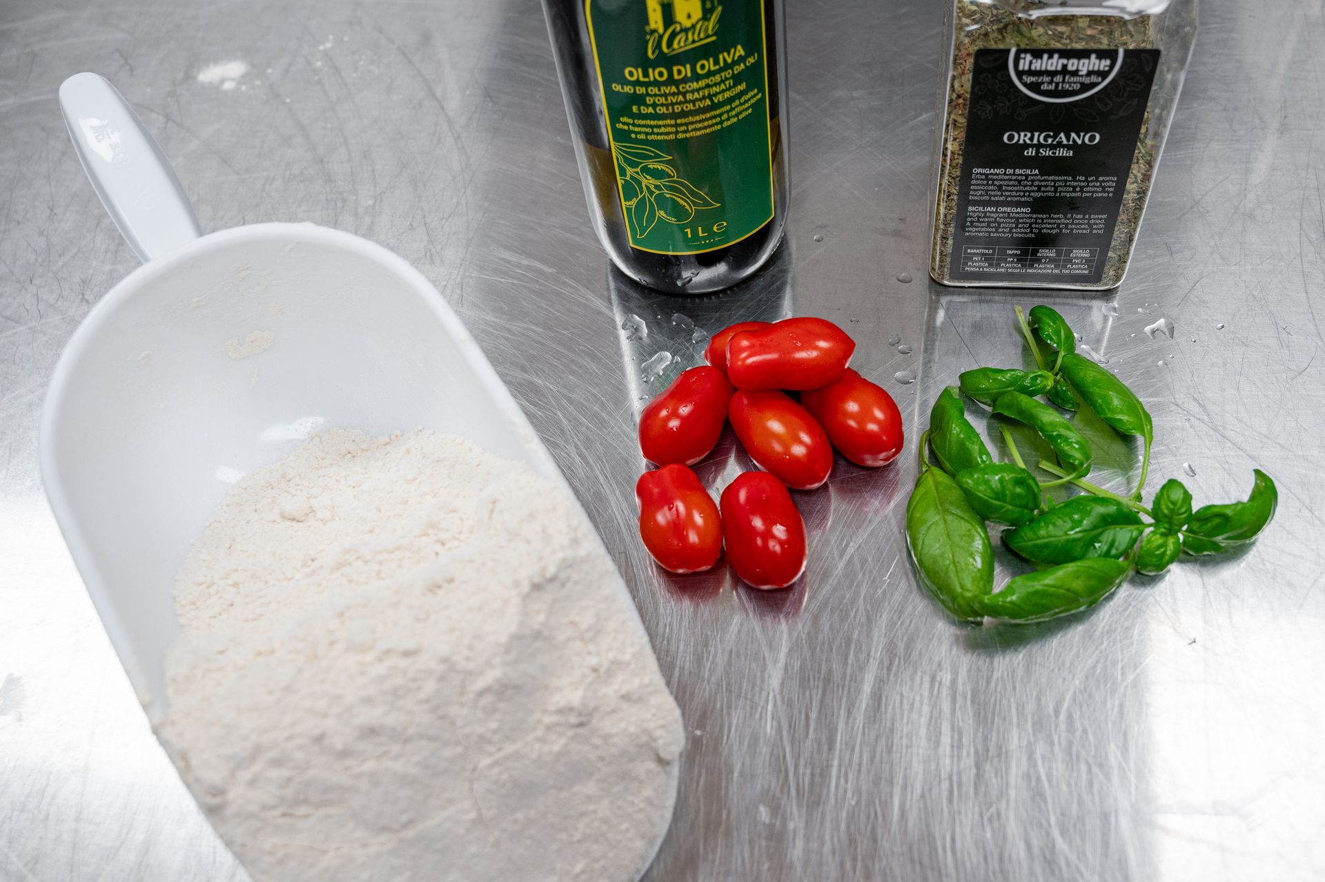 Preparazione degli ingredienti con farina, pomodorini, foglie di basilico e olio d'oliva su un piano di lavoro in metallo.