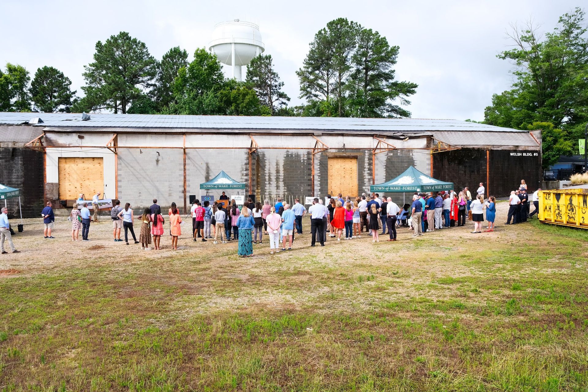 Group of people gathered outside a building with boarded-up doors; tents are set up. Trees and water tower in background.
