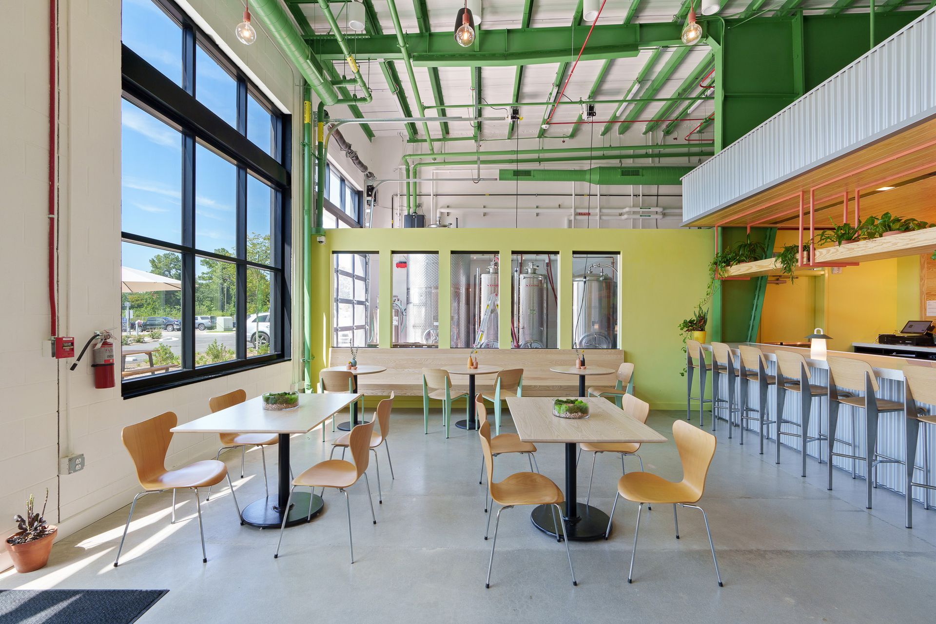 Bright, modern brewery interior. Tables and chairs in front of large windows, bar in the background. 