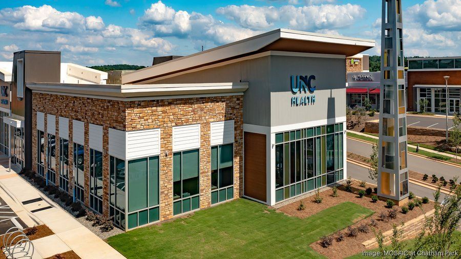 UNC building exterior. Light blue and stone facade with large windows, green lawn, and blue sky.