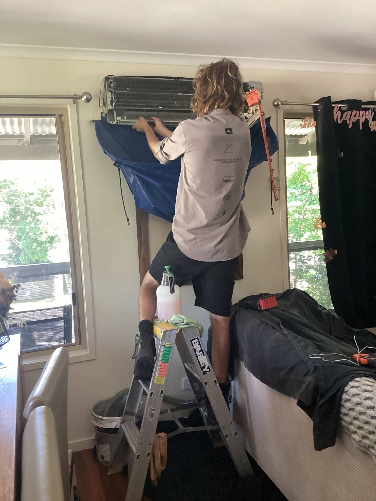 A Man is Standing on a Ladder Cleaning an Air Conditioner — Kilowatts & Cool Whitsundays In Strathdickie, QLD