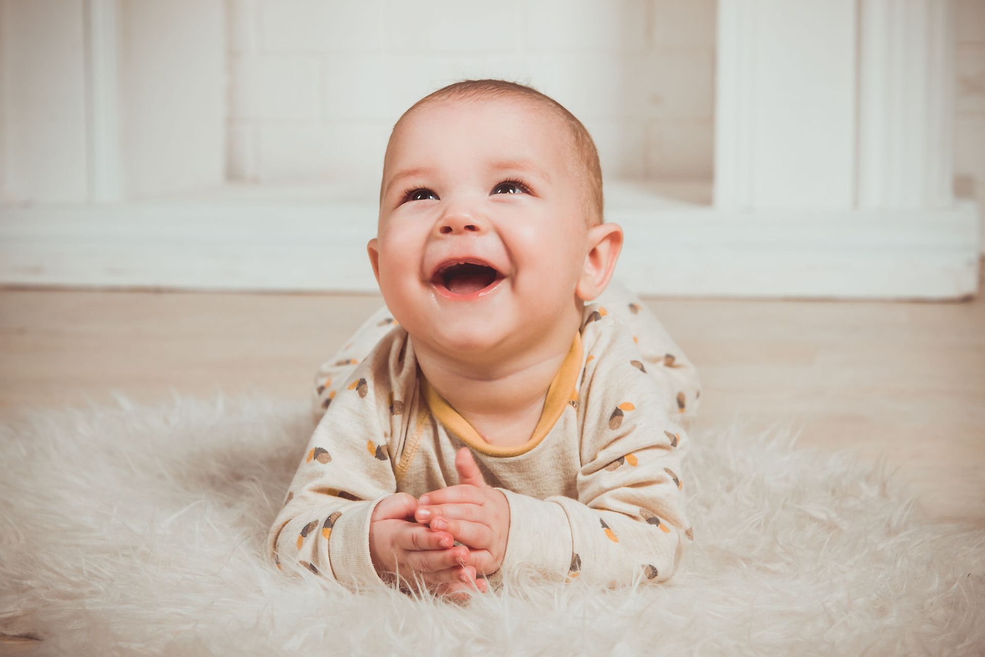 Un bebé sonriente yace sobre una alfombra mullida en el interior de la casa, con las manos entrelazadas y mirando hacia arriba.