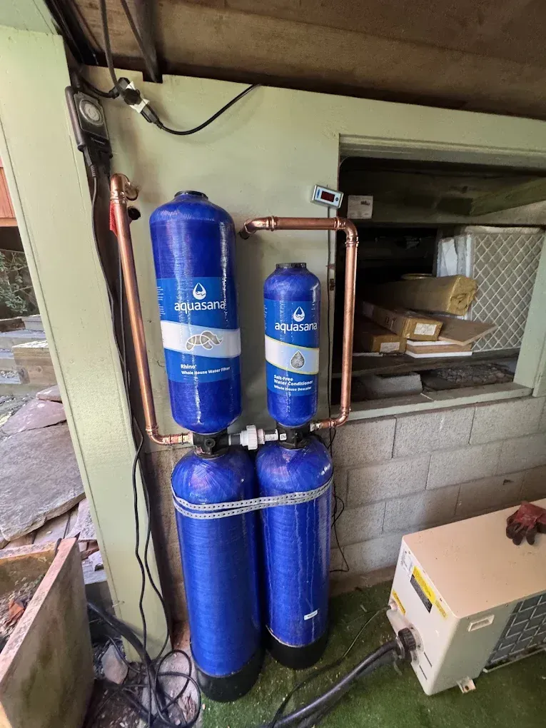 Four blue water filtration tanks and copper pipes installed beside a cinder block wall in a utility area