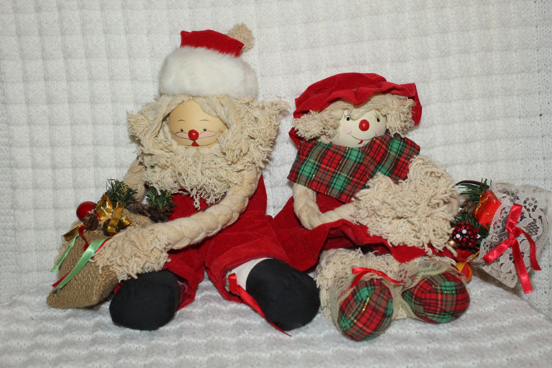Two Santa Claus plush dolls with gift bags, sitting on a white textured blanket