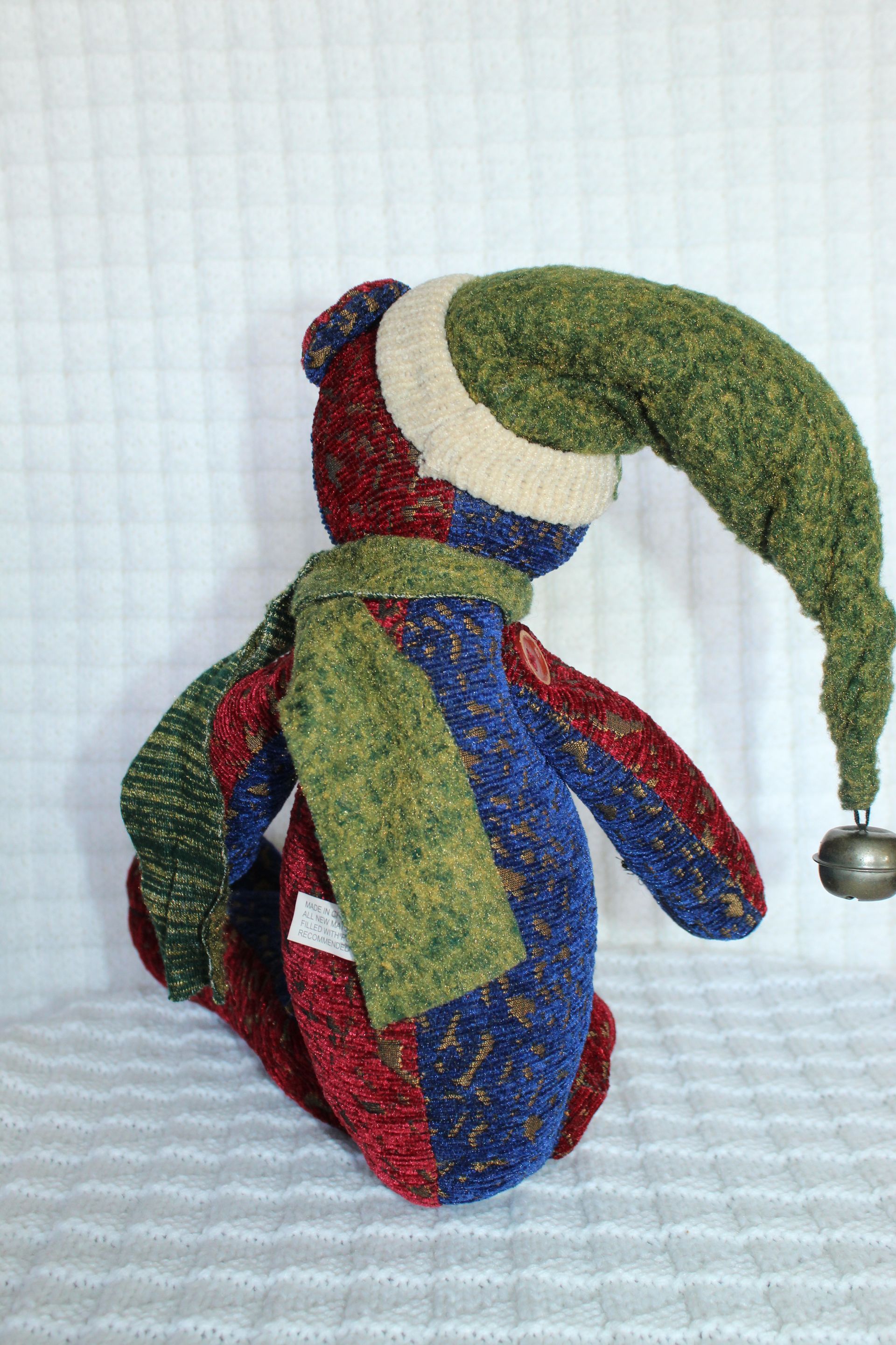 Colorful knitted jester doll with green hat, blue-red body, and dangling bell, sitting on a white surface.