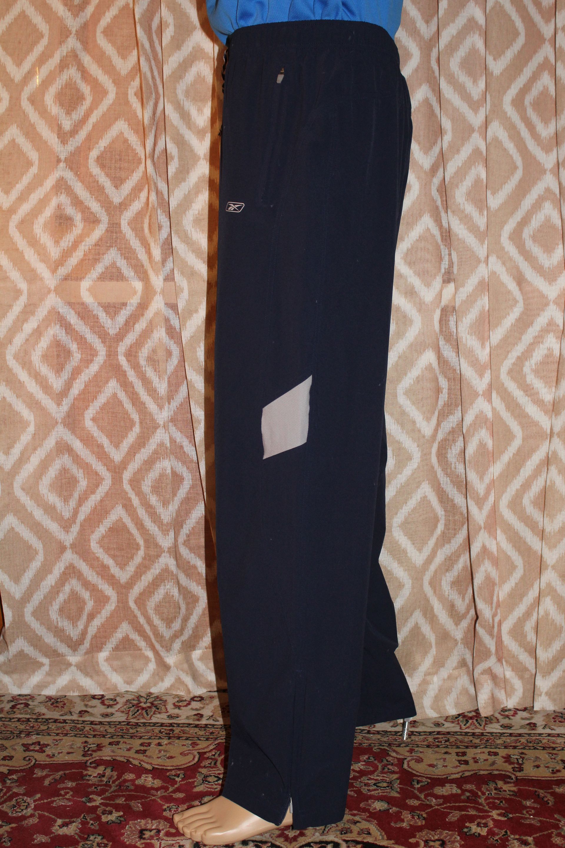 Person standing indoors wearing a blue top and black pants against a patterned curtain.