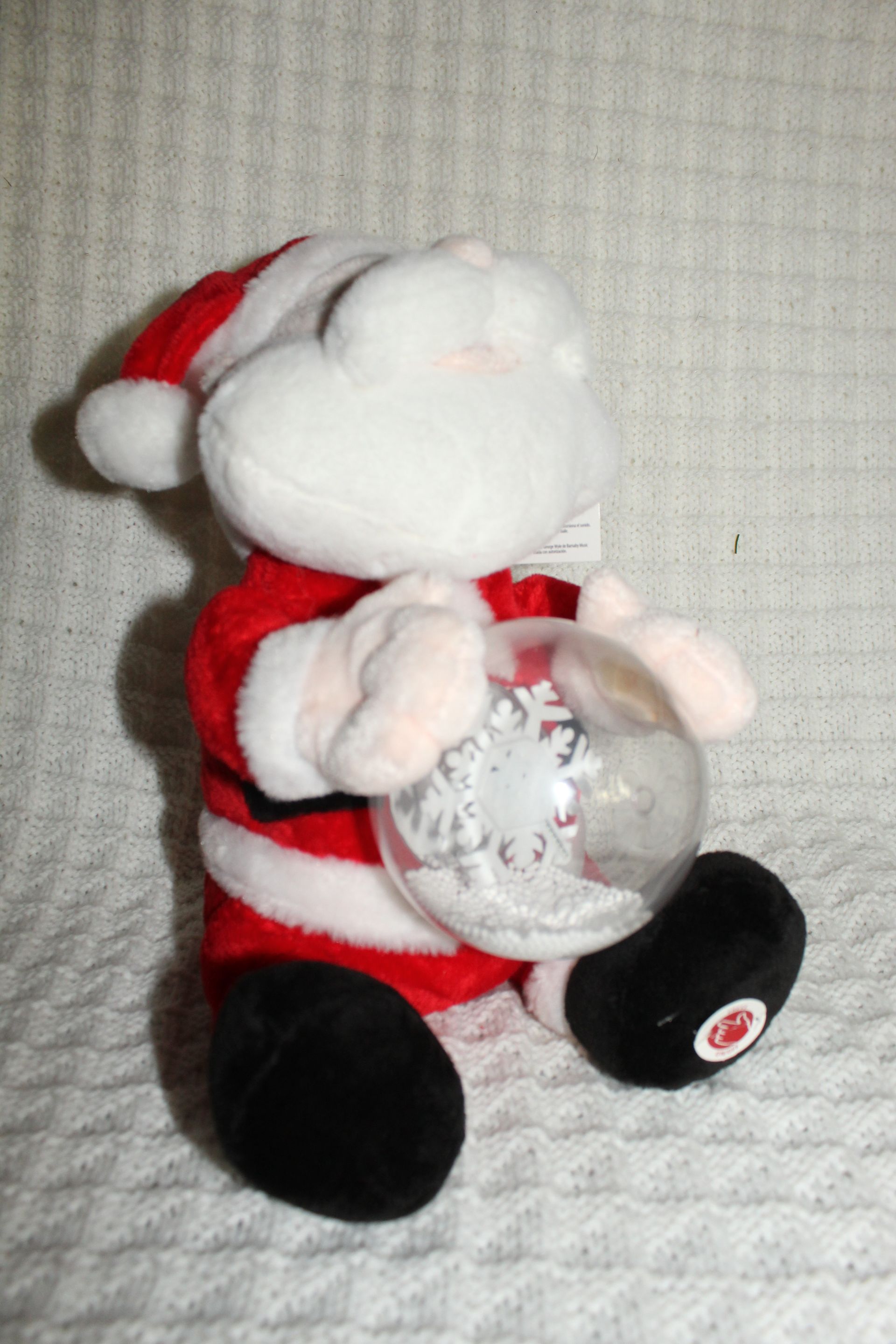 Plush Santa figurine holding a clear ornament, wearing a red suit and black boots against a white textured background