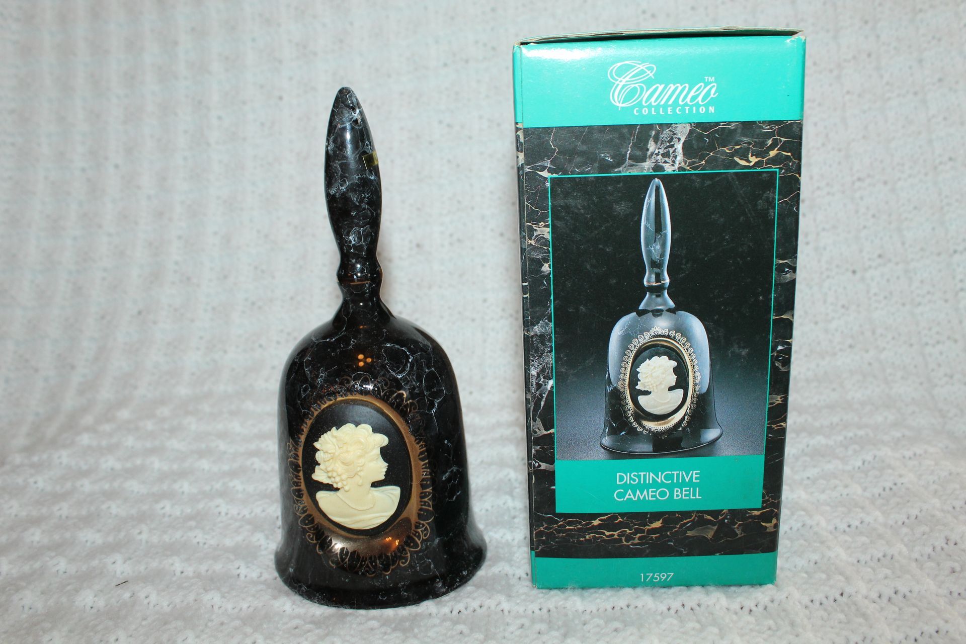 Black decorative bell with cameo portrait on a white textured surface, shadow cast behind it