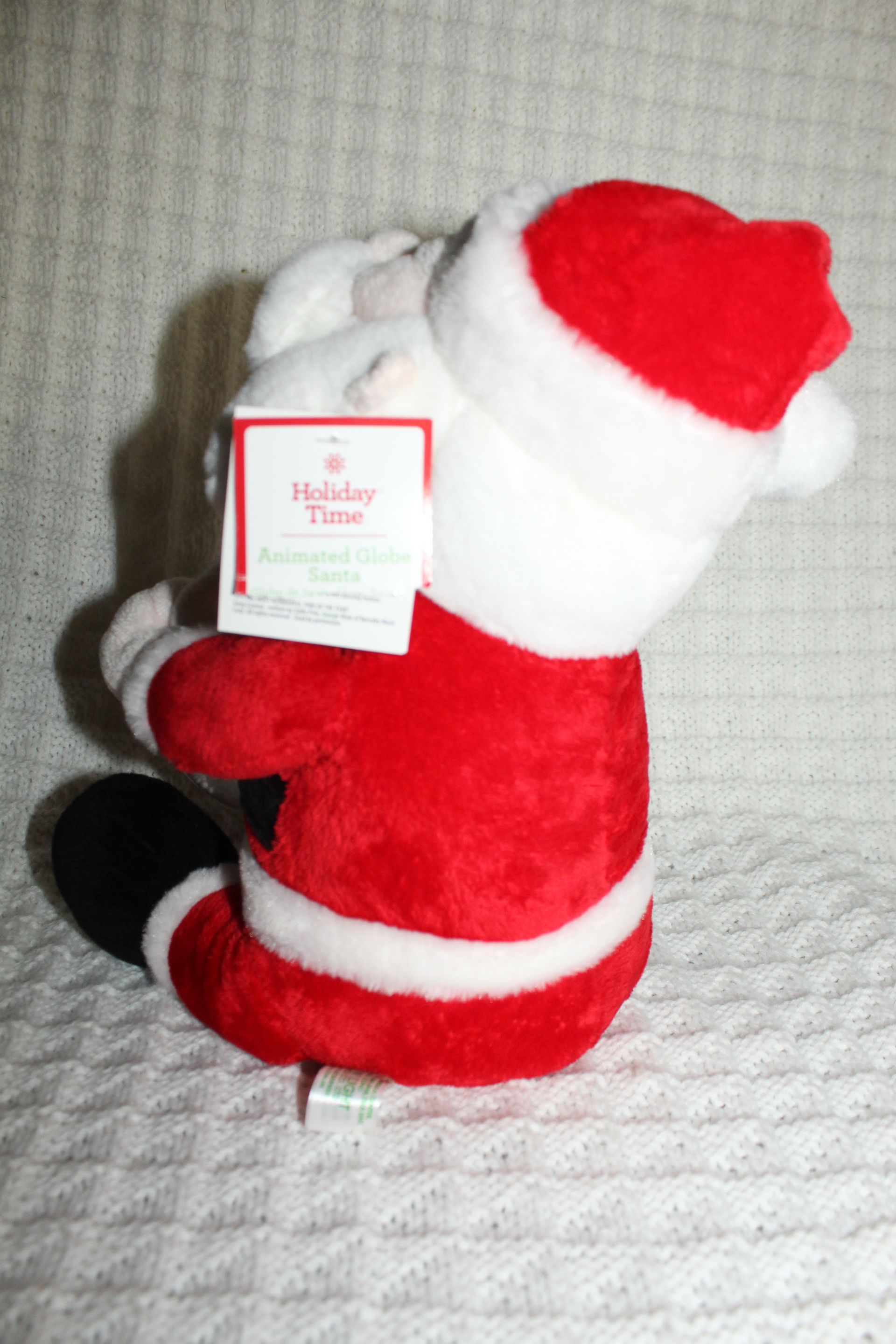 Plush Santa figurine holding a clear ornament, wearing a red suit and black boots against a white textured background