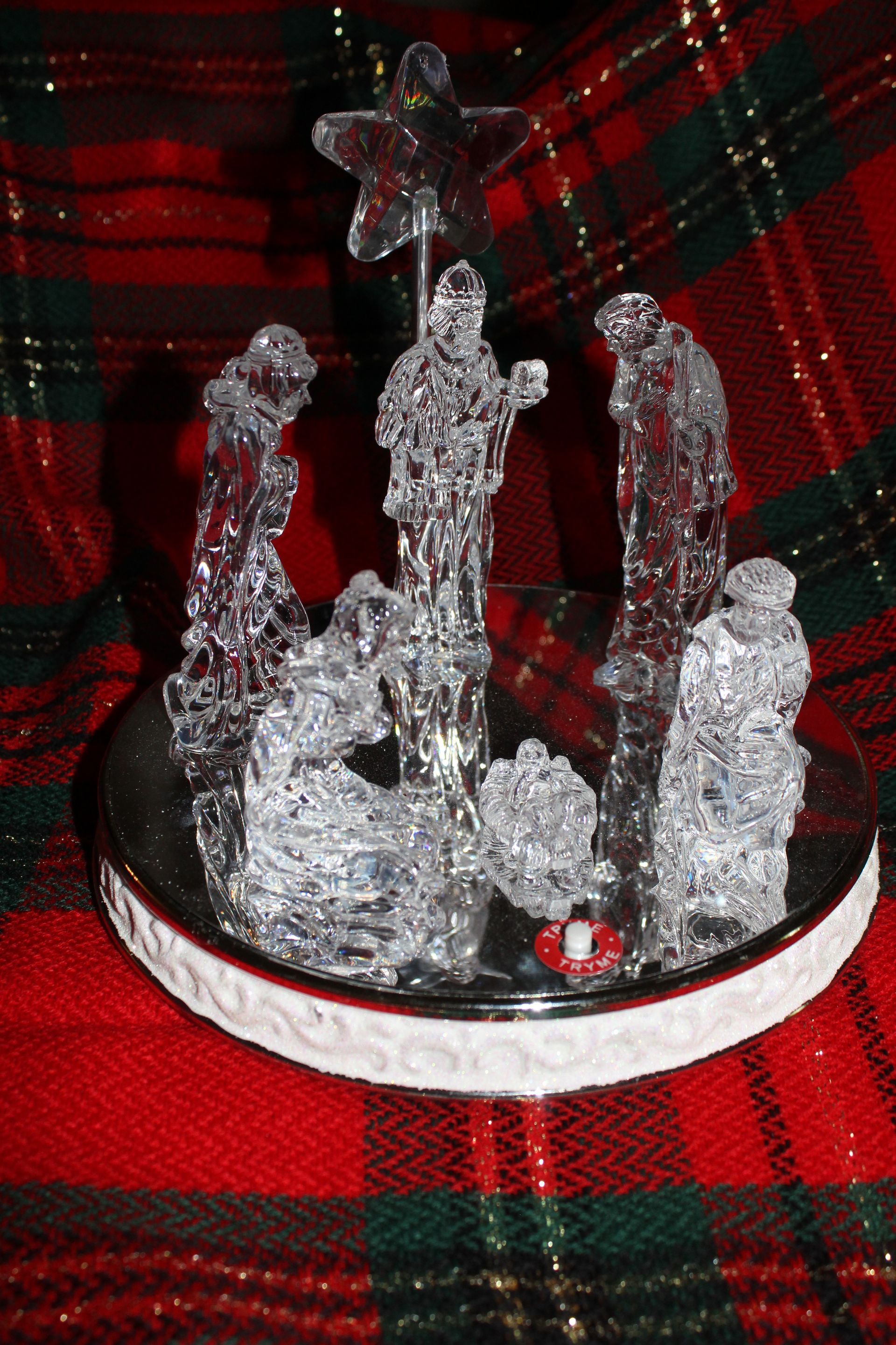Small clear nativity figurines on a round white base against red-and-green plaid fabric.